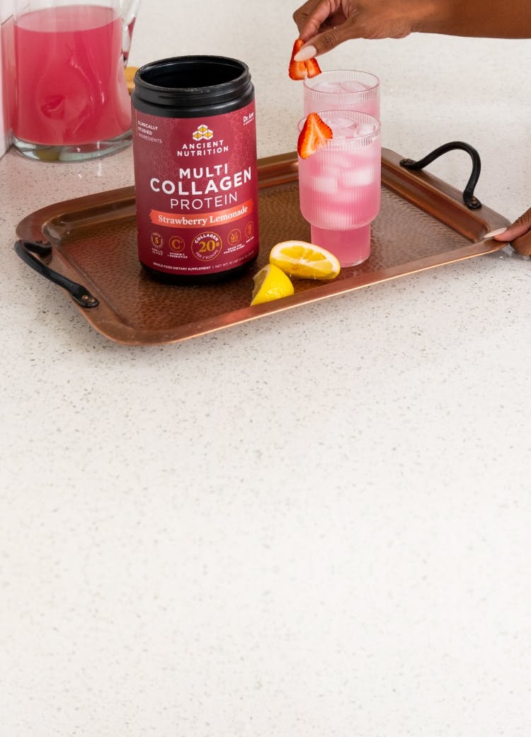 A container of Ancient Nutrition Multi Collagen Protein Strawberry Lemonade on a bronze on a counter with a glass of it mixed in water with ice.