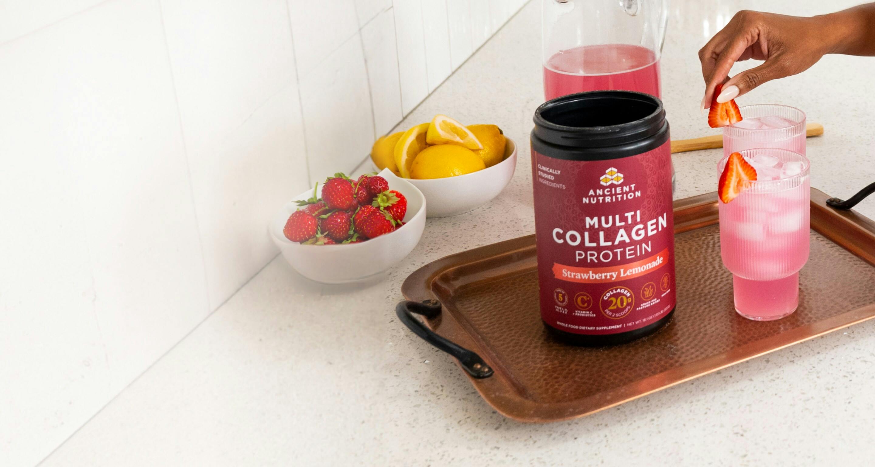 A container of Ancient Nutrition Multi Collagen Protein Strawberry Lemonade on a counter with a glass of it mixed in water with ice and a woman's hand.