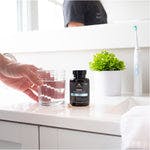 a man picking up a glass of water on a bathroom counter next to a bottle of  SBO Probiotics Men's capsules