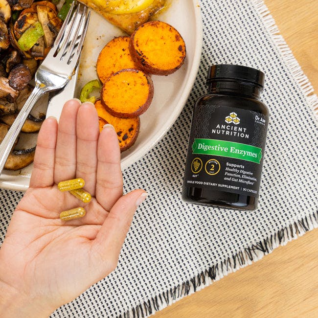person holding digestive enzyme capsules in their hand next to plate of veggies