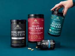 Bottles of Ancient Nutrition bone broth protein, multi collagen protein, organic supergreens, and sbo probiotics on teal background with a woman's hand reaching for one